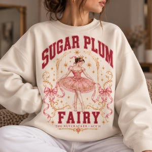 Peut inclure: Un sweat-shirt à col rond couleur crème avec une impression graphique représentant une ballerine en tutu rose. Le texte indique SUGAR PLUM FAIRY et THE NUTCRACKER ACT II en lettres rouges, entouré de filigranes dorés, de rubans roses et d'étoiles.