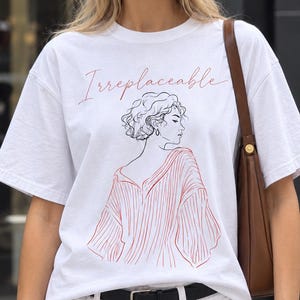 May include: A white short-sleeved t-shirt featuring a red line-art illustration of a person with curly hair wearing a striped top. The word Irreplaceable is printed in red cursive script above the illustration. The shirt has a crew neckline.