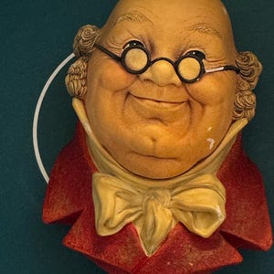May include: A wall-mounted decorative plaque featuring a sculpted bust of a bald, smiling character wearing round black spectacles, a cream-colored cravat, and a textured red coat. The figure has a light tan complexion and curly hair at the sides.