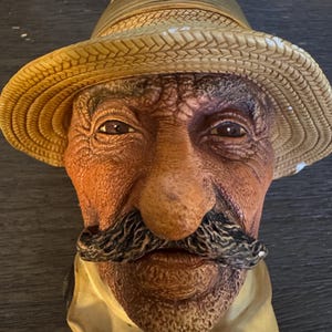 May include: A sculpted bust of an older individual with a textured, weathered complexion, a prominent dark mustache, and deep-set eyes. The figure wears a tan straw-textured hat with a wide brim and a light-colored collared shirt underneath.