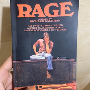 Könnte beinhalten: Ein Taschenbuch mit dem Titel Rage von Richard Bachman. Das schwarze Cover zeigt einen großen orangefarbenen Schriftzug, eine Person, die auf einem Schreibtisch sitzt und das Kinn auf eine Hand stützt, sowie Beine in roter Kleidung am Boden.
