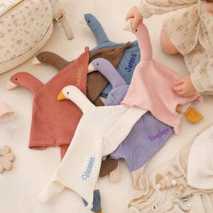 Puede incluir: Una colección de mantitas de seguridad de muselina con forma de ganso en varios colores, incluyendo rosa, azul, marrón, morado y crema. Cada mantita presenta un nombre bordado como Charlotte, James, Penelope y Victoria en la tela.