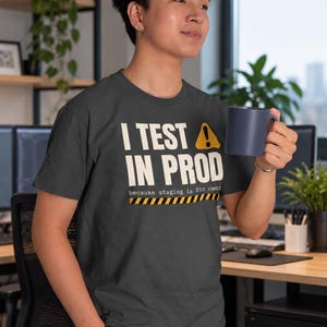 以下が含まれることがあります： ダークグレーの半袖Tシャツ。白い文字で「I TEST IN PROD」、黄色の警告三角形アイコン、その下に「because staging is for cowards」という文字、さらにその下に黄色と黒の危険警告ストライプ模様がプリントされています。