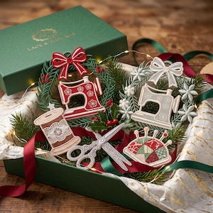 May include: A gift box containing embroidered sewing-themed ornaments including a red sewing machine in a wreath, a light gray sewing machine in a floral wreath, a brown thread spool, silver scissors, and a colorful pincushion with the text LACE & THREADS.