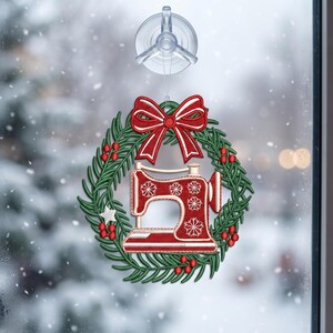 May include: A decorative hanging ornament featuring a red sewing machine with white snowflake patterns, centered within a green pine wreath adorned with red berries and a white star, topped with a red bow, suspended by a clear suction cup.