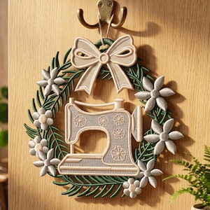May include: A decorative wall hanging featuring a green wreath adorned with light beige flowers and a matching bow at the top. Centered within the wreath is a light beige sewing machine silhouette with a detailed floral pattern on its side.
