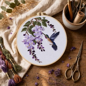 Op de afbeelding: Een ronde houten borduurring met witte stof en een handgeborduurd ontwerp van een kolibrie die bij een tros paarse blauweregenbloemen met groene bladeren zweeft. Het borduurwerk is omringd door borduurgaren en een schaar.