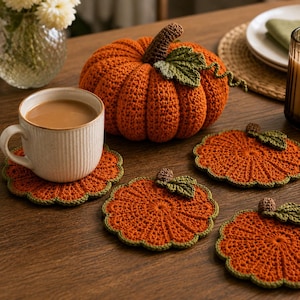 May include: A set of orange crochet pumpkin coasters with green leaves and brown stems, accompanied by a larger matching crochet pumpkin and a white ceramic mug filled with a light brown beverage, all arranged on a wooden surface.