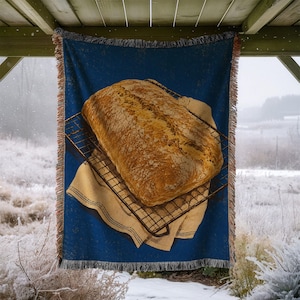 May include: A woven throw blanket featuring a deep blue background with a central graphic of a golden-brown loaf of bread resting on a black wire cooling rack atop a tan cloth, finished with fringed edges on the top and bottom sides.
