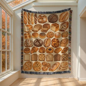 May include: A woven throw blanket featuring a grid of various bread types, including baguettes, sourdough loaves, sliced sandwich bread, croissants, and rolls, all rendered in shades of brown, tan, and cream with a fringed edge.