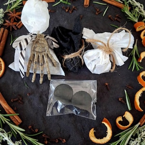 Könnte beinhalten: Drei kleine Stoffbündel, die mit Bindfaden zusammengebunden sind, eines davon mit einer Skeletthand aus Kunststoff und schwarz-weiß gestreiftem Band. Ein durchsichtiger Plastikbeutel enthält zwei runde schwarze Gegenstände. Das Arrangement ist umgeben von Rosmarin, Zimtstangen, Sternanis, Nelken und Orangenschalen.