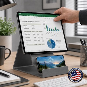 May include: A dark gray metal dual-device stand holding a tablet displaying a Q2 Marketing Budget spreadsheet with bar and pie charts, and a smartphone showing a mountain landscape wallpaper with the time 11:41 on the lock screen.