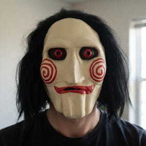 May include: A full-face mask featuring a pale off-white complexion, prominent red spiral patterns on the cheeks, and deep-set red eyes. The mask includes a sculpted red mouth area and is framed by thick, straight black hair attached to the sides.