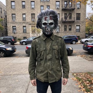 May include: A full-head latex mask featuring a grey, weathered zombie face with dark eye sockets, exposed teeth, and messy grey hair, paired with an olive green military-style field jacket with four front pockets and button-down shoulder epaulets.