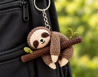 May include: A crocheted sloth keychain featuring a brown sloth with a cream-colored face, pink cheeks, and dark brown eye patches, hanging from a brown branch with small green leaves, attached to a silver-toned metal keyring and chain.