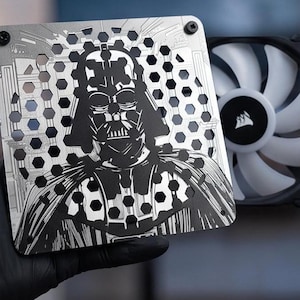 May include: A square silver metal computer fan grille featuring a laser-cut design of Darth Vader. The grille has a repeating hexagonal pattern throughout the surface and is positioned in front of a black computer cooling fan with white blades.