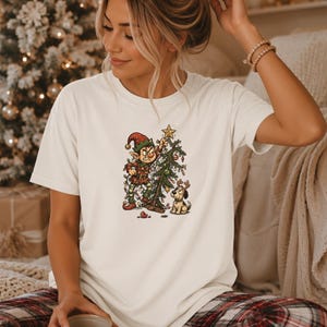 Peut inclure: Un t-shirt à manches courtes de couleur crème avec une impression graphique représentant un elfe malicieux emmêlé dans des guirlandes lumineuses de Noël, plaçant une étoile sur un petit sapin, avec un petit chien portant des bois de renne, et le texte NATURAL.