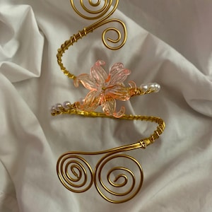 Peut inclure: Un bracelet de bras en fil métallique doré avec un design en spirale aux deux extrémités. Le fil est enroulé en spirale et orné d'une fleur translucide rose au centre, entourée de petites perles sphériques blanches de chaque côté.