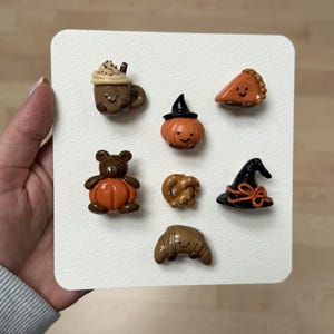 May include: A set of seven small, glossy, sculpted clay charms arranged on a white square card. The charms include a brown mug with whipped cream, a pumpkin wearing a black witch hat, a slice of pumpkin pie, a bear holding a pumpkin, a pretzel, a black witch hat with an orange ribbon, and a brown croissant.