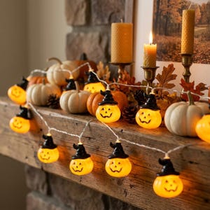 Puede incluir: Una cadena de luces LED con pequeñas calabazas naranjas que llevan sombreros de bruja negros. Las figuras de calabaza brillantes están conectadas por un cable blanco fino, intercaladas con varias calabazas decorativas blancas y naranjas y piñas.
