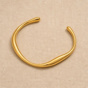 Peut inclure: Un bracelet manchette en métal doré au design ouvert avec une finition lisse et polie. Le bracelet présente une forme organique irrégulière avec des extrémités arrondies et effilées, créant une silhouette fluide et continue en cercle.
