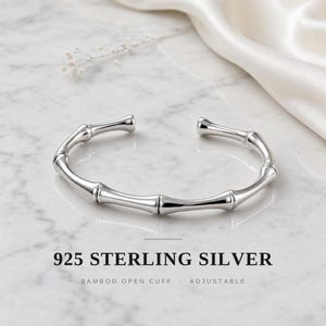 Puede incluir: Una pulsera abierta de plata de ley 925 pulida, diseñada con una textura segmentada de tallo de bambú. La pieza de joyería es ajustable y presenta un acabado metálico liso y reflectante. El texto indica 925 STERLING SILVER BAMBOO OPEN CUFF ADJUSTABLE.