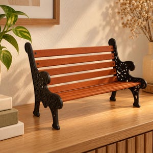 May include: A miniature park bench featuring a backrest and seat made of horizontal brown wooden slats. The frame and armrests are constructed from black metal with intricate, ornate scrollwork patterns visible on the side supports.