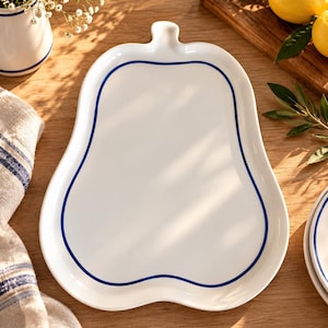 Könnte beinhalten: Eine weiße Servierplatte aus Keramik in Birnenform mit glatter Oberfläche und einer einzelnen dünnen dunkelblauen Linie, die den Umriss der Fruchtform nachzeichnet. Die Platte hat ein kleines Stiel-Detail oben und einen abgerundeten Boden.