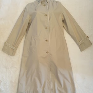 May include: A beige trench coat featuring a high collar with a buckle strap, a front button closure, and long sleeves with adjustable buckled cuffs. The coat has a smooth finish and a clean, structured design with a white trim along the front placket.