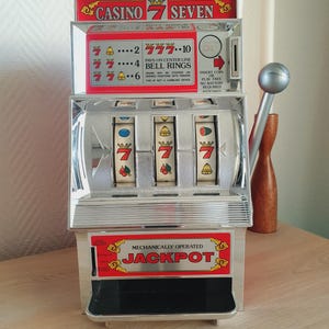 Könnte beinhalten: Ein silberfarbenes mechanisches Spielautomat-Spielzeug mit roten Akzenten, einem seitlichen Hebel und drei rotierenden Walzen mit Symbolen wie Siebenen, Glocken und Kirschen. Das Gerät zeigt den Text CASINO 7 SEVEN und MECHANICALLY OPERATED JACKPOT.