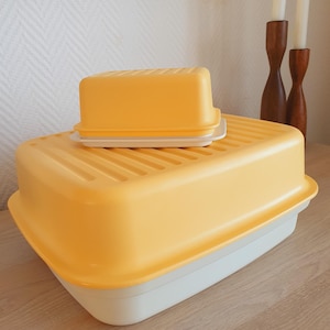 Op de afbeelding: Een rechthoekige gele en witte plastic botervloot met een geribbeld deksel staat op een grotere rechthoekige gele en witte plastic opbergdoos met een bijpassend geribbeld deksel. Beide containers hebben een witte basis en een gele deksel.