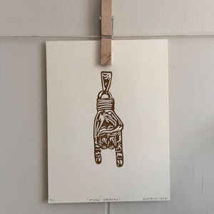 May include: A brown linocut print of a hand making the 'rock on' gesture, hanging from a wooden clothespin. The print is on off-white paper and includes the text "MANO LOCAUTO" and "ALEXAOS 2019".