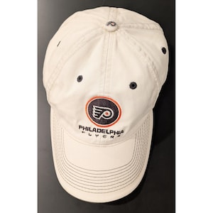 Op de afbeelding: Een crèmekleurige baseballpet met het Philadelphia Flyers-logo in zwart en oranje op de voorzijde geborduurd. De pet is voorzien van donkerblauwe oogjes, een gebogen klep met decoratieve stiksels en een klein logo op de bovenste knoop.
