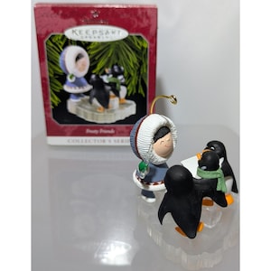Könnte beinhalten: Ein Hallmark Keepsake Ornament mit dem Titel Frosty Friends, das eine Figur in einem blauen Parka mit weißer Fellkapuze auf einem transparenten Sockel zeigt, zusammen mit drei schwarz-weißen Pinguinen, von denen einer einen grünen Schal trägt.