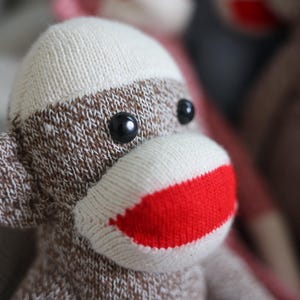 Puede incluir: Un peluche de mono hecho de tela de punto jaspeada en marrón y blanco. El juguete presenta una cara de color crema con una boca roja brillante, dos pequeños ojos de botón negros y orejas redondeadas que sobresalen a los lados de la cabeza.