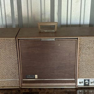 May include: A portable Zenith Stereophonic record player with a brown wood-grain finish, a top carrying handle, and two integrated speakers featuring a tan woven mesh grille. The front panel displays the Zenith Stereophonic logo and four silver control knobs.