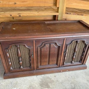 May include: A rectangular wooden console cabinet featuring a dark brown finish. The front displays three sections: two outer panels with vertical fabric speaker grilles flanked by turned wooden spindles, and a central solid wood panel door.