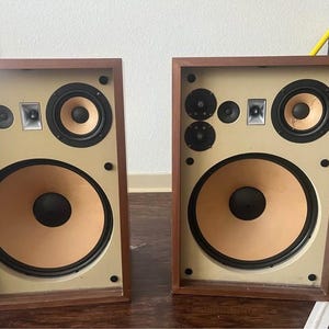 May include: A pair of rectangular wooden speakers with a light beige front panel. Each speaker features one large woofer with a black centre, one mid-range driver, two small tweeters, and a rectangular silver-toned port, all framed in dark wood.