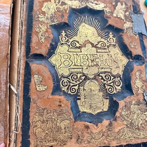 Puede incluir: Un libro antiguo con una cubierta de cuero marrón y negro desgastada que presenta ilustraciones doradas en relieve y la palabra BIBELN centrada en la parte frontal. La cubierta muestra escenas religiosas, bordes decorativos y cierres.