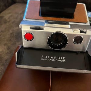 May include: A vintage Polaroid SX-70 Land Camera featuring a brushed silver metal body with brown leather accents. The front includes a bright red shutter button, a circular lens with a black dial, and a black panel labeled POLAROID SX-70 LAND CAMERA.