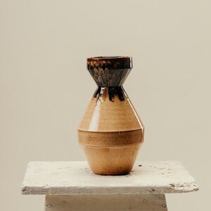 Può includere: Un vaso in ceramica con un corpo ampio e angolare e un collo stretto. La parte inferiore è di colore marrone chiaro, mentre il collo superiore e il bordo presentano una smaltatura lucida marrone scuro che cola, creando un netto contrasto.