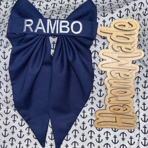 May include: A navy blue fabric bow with the name RAMBO embroidered in white thread across the center band. The bow is positioned next to a light-colored wooden cutout sign with the word HeroMade written in a cursive script font.
