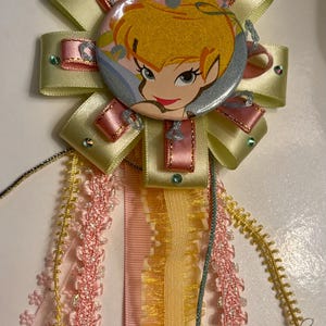 May include: A decorative rosette badge featuring a central circular button with a cartoon character illustration of a blonde fairy. The badge is layered with pale yellow and pink satin ribbons, accented with small rhinestones and hanging lace trims.