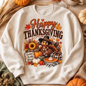 Può includere: Una felpa girocollo color crema con una stampa grafica di un tacchino con cappello e sciarpa, circondato da zucche, un girasole, foglie autunnali e una torta. Il testo recita Happy Thanksgiving e Grateful Thankful Blessed.
