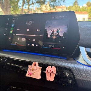 May include: A light pink miniature handbag and a pair of matching light pink slide sandals attached to a car air vent. The handbag features a small gold-tone clasp and a tiny pink charm. The car dashboard displays a digital media interface.
