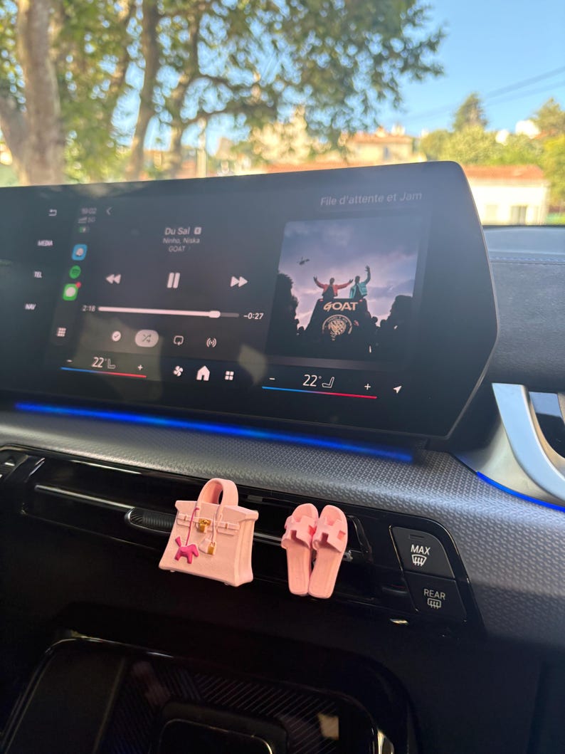 May include: A light pink miniature handbag and a pair of matching light pink slide sandals attached to a car air vent. The handbag features a small gold-tone clasp and a tiny pink charm. The car dashboard displays a digital media interface.
