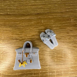 Op de afbeelding: Een lichtgrijze handtasbedel met een miniatuur gestructureerde tas met goudkleurige hardware en een kleine gele paardvormige hanger, vergezeld van een paar lichtgrijze slippers met het woord HERMES in reliëf op het voetbed.