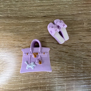 Peut inclure: Un sac à main lavande avec un fermoir doré, une petite breloque cheval blanche et un cadenas doré assorti, accompagné d'une paire de sandales à enfiler lavande présentant un motif de bride en forme de H découpé sur le dessus.