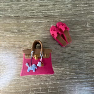 Op de afbeelding: Een miniatuur roze handtas met bruine hengsels, een goudkleurig slotje en een lichtblauw paardenbedeltje, vergezeld door een paar felroze slippers met de tekst HERMES op de bruine binnenzolen, alles uitgevoerd als kleine decoratieve items.