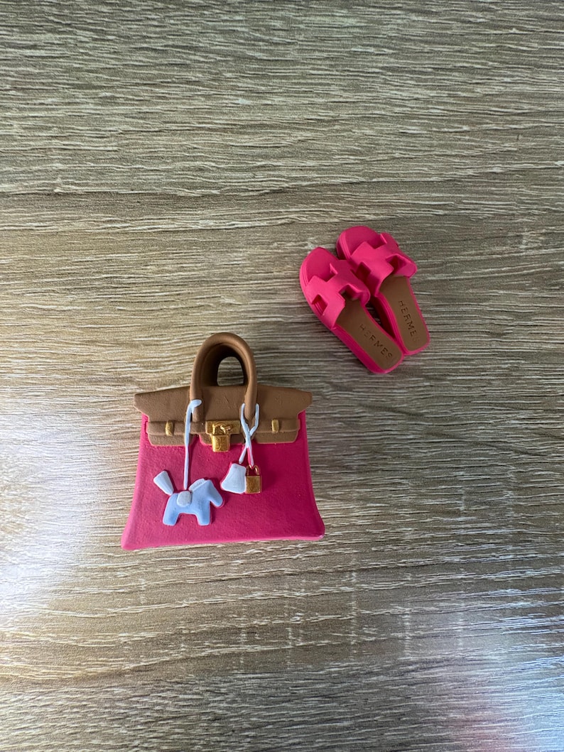 May include: A miniature pink handbag with brown handles, a gold-tone lock, and a light blue horse charm, accompanied by a pair of bright pink slide sandals featuring the text HERMES on the brown insoles, all crafted as small decorative items.